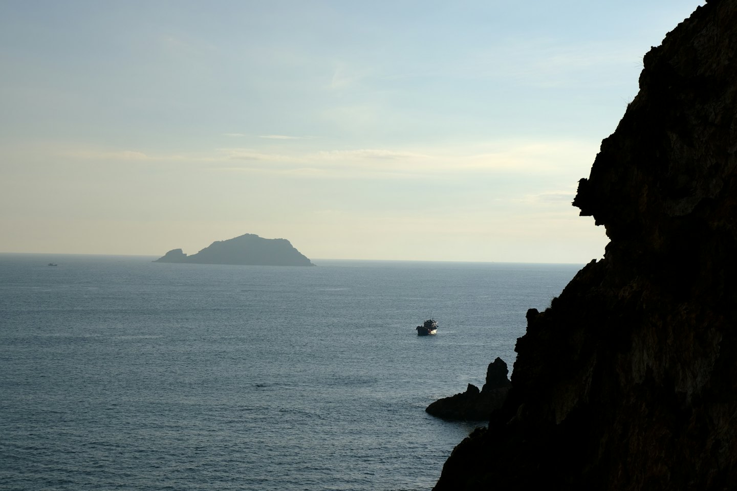 A fishing boat sails on calm blue waters near a cliff.
