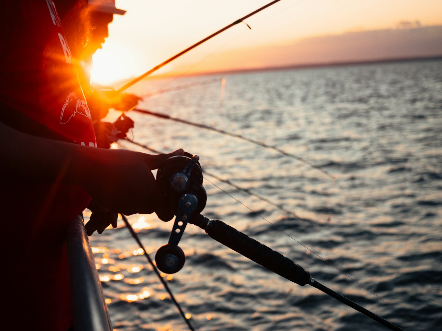 Hands operate a fishing reel on a boat at sunset.