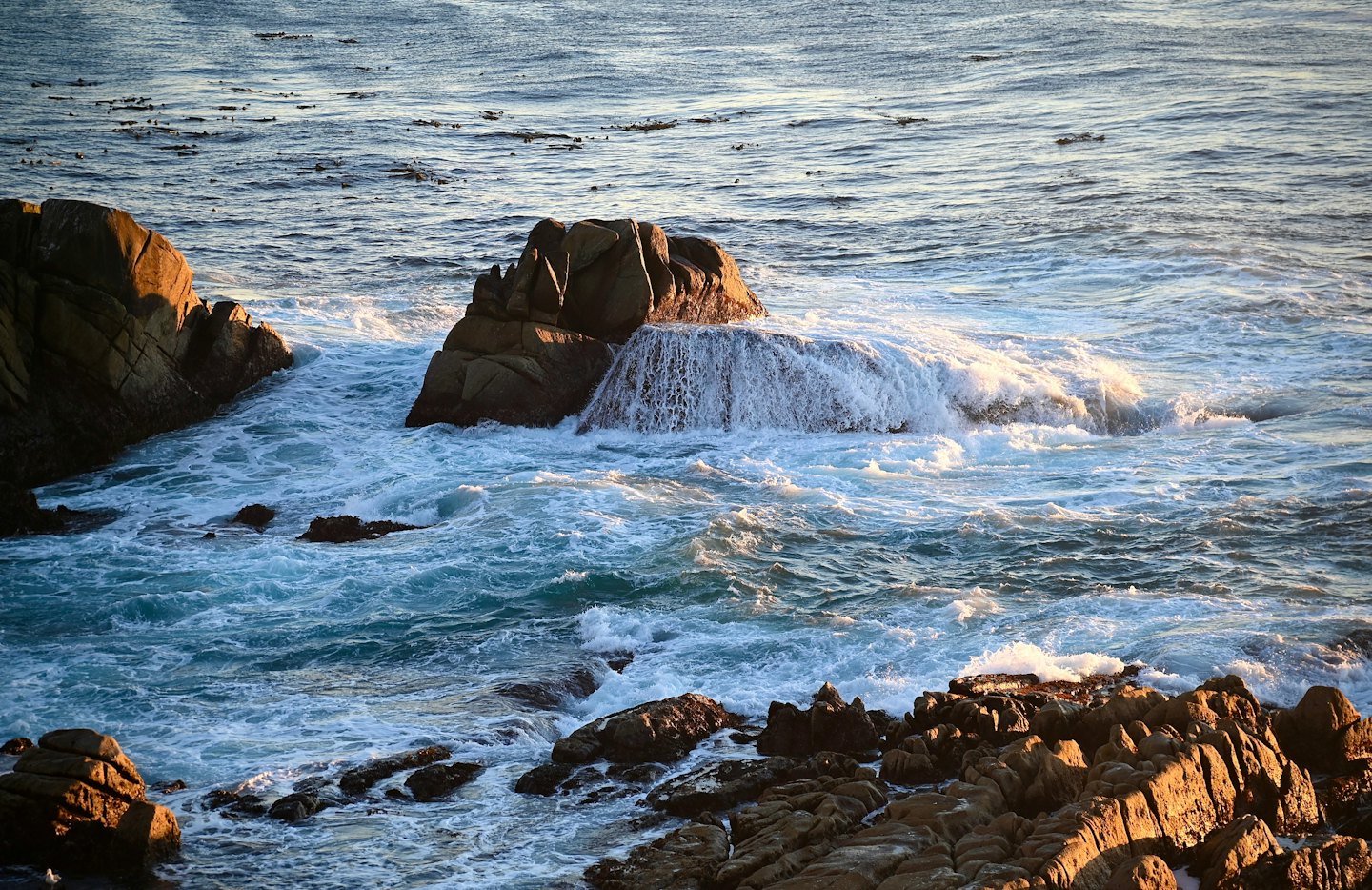 Ocean waves crashing on dark, jagged rocks at sunset.